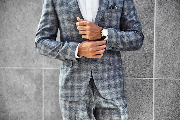 Modelo de empresario de moda vestido con elegante traje a cuadros posando junto a la pared gris en el fondo de la calle. Metrosexual con reloj de lujo en muñeca