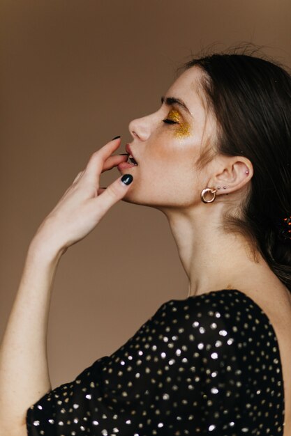 Modelo europea sensual lleva accesorios dorados de pie sobre una pared marrón. Foto interior de una encantadora mujer de pelo negro con maquillaje de fiesta brillante.