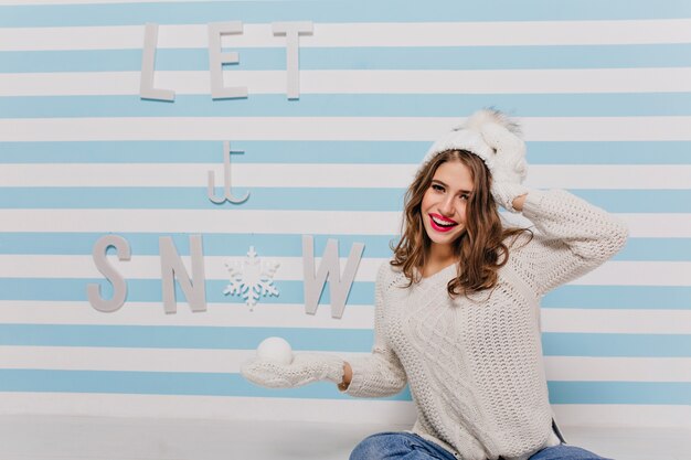 Modelo feliz y encantador con piel delicada y adorable sonrisa linda posando en ropa de invierno, sentada contra la pared con hermosa inscripción