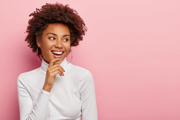 La modelo femenina despreocupada satisfecha sonríe suavemente, toca la barbilla, mira a un lado, nota una escena divertida, se ríe de algo, tiene el cabello oscuro y rizado natural, se viste informalmente, aislado en una pared rosa