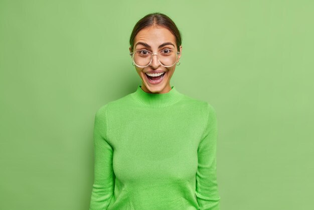 La modelo femenina europea alegre positiva se siente sorprendida ríe alegremente usa lentes transparentes y pses de cuello alto contra la pared verde vivo