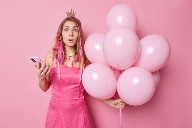 Una modelo femenina impresionada y atónita tiene la imagen de una princesa que lleva una pequeña corona y un vestido sostiene un teléfono móvil recibe un mensaje inesperado de un novio formal que viene a la fiesta con un montón de globos inflados