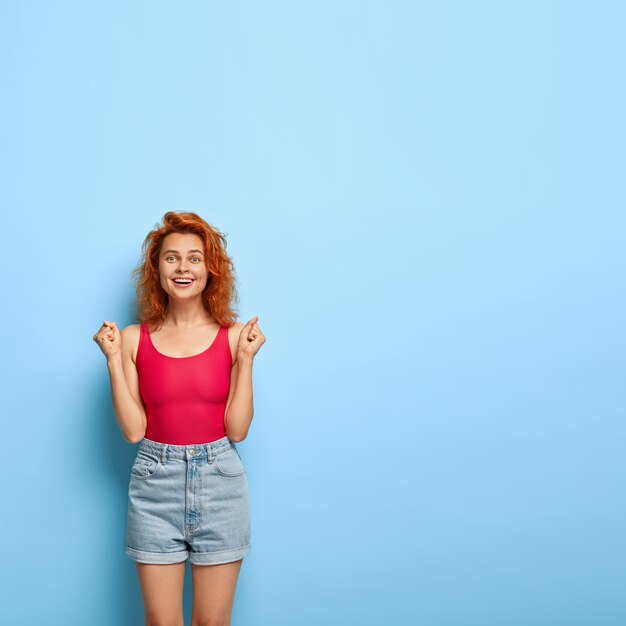 La modelo femenina de jengibre complacida aprieta los puños, anticipa algo bueno, sonríe alegremente, usa chaleco rojo y pantalones cortos de mezclilla