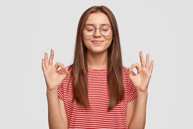 La modelo femenina joven satisfecha y complacida no hace ningún gesto, usa gafas transparentes, tiene el pelo largo y oscuro