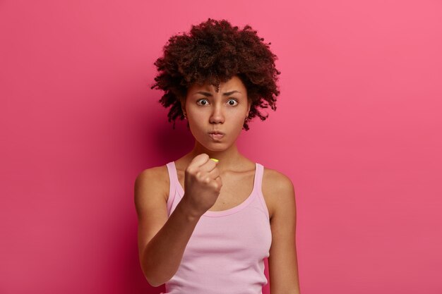 La modelo femenina de piel oscura e irritada feroz aprieta el puño con ira, demuestra fuerza y poder, está molesta con alguien, usa ropa informal, se para contra la pared rosa vibrante.