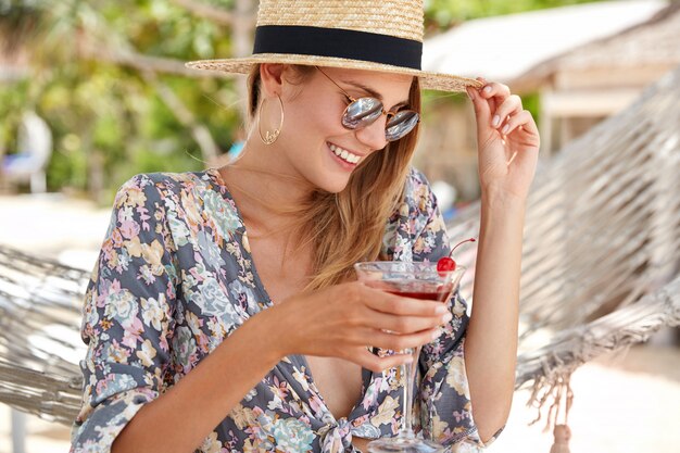 La modelo femenina relajada y contenta se siente emocionada mientras se recrea al aire libre en un país tropical cálido, usa una camisa estampada de flores y un sombrero de paja, gafas de sol de moda, bebe un cóctel de cereza fresca,