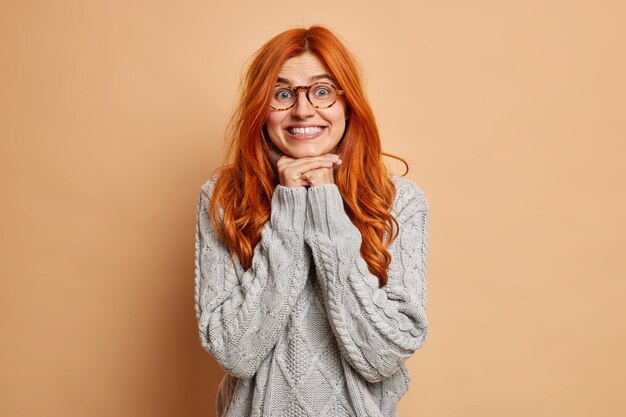 La modelo femenina satisfecha mantiene las manos bajo la barbilla se ve con una expresión curiosa y sonríe ampliamente vestida con un suéter de punto.