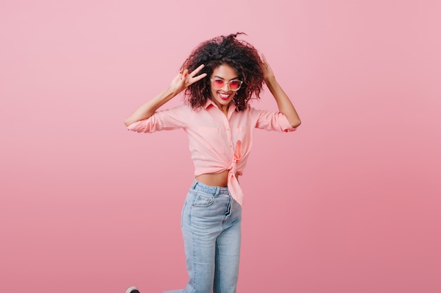 Foto gratuita modelo femenino africano delgado en pantalones de mezclilla vintage que expresan felicidad. glamorosa chica bien formada en ropa casual riendo cerca de la pared rosa.