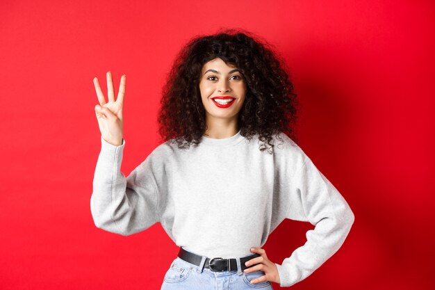 Modelo femenino alegre que muestra el número tres y sonriendo, haciendo un pedido, de pie en una sudadera sobre fondo rojo.
