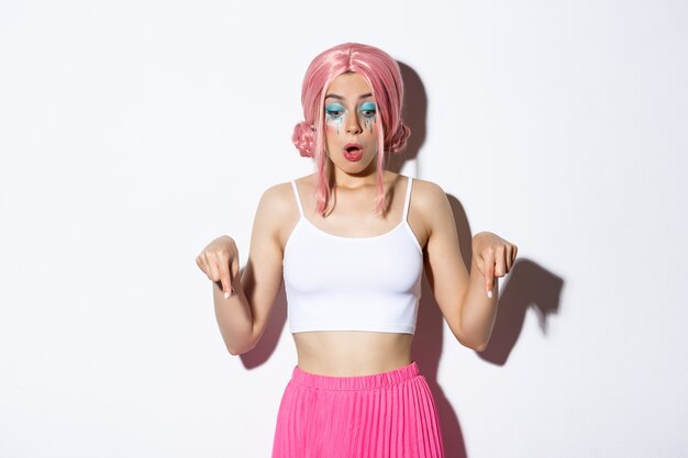 Modelo femenino asombrado con peluca rosa y maquillaje brillante, jadeando de asombro, señalando con el dedo hacia abajo, mirando el logotipo o la pancarta, blanco.