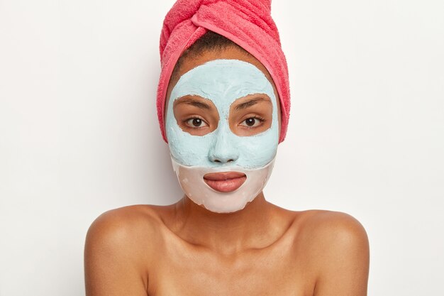 Modelo femenino de aspecto agradable con piel fresca, aplica mascarilla de belleza, usa una toalla rosa en la cabeza, se para con los hombros desnudos, mira directamente