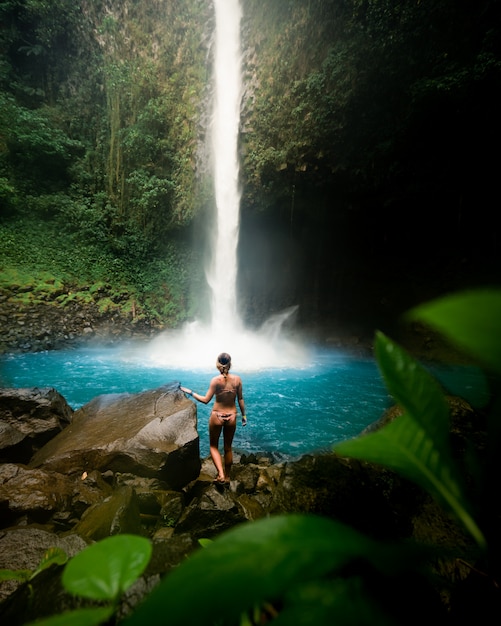 Modelo femenino atractivo en bikini de pie sobre las rocas cerca de una hermosa cascada en un bosque