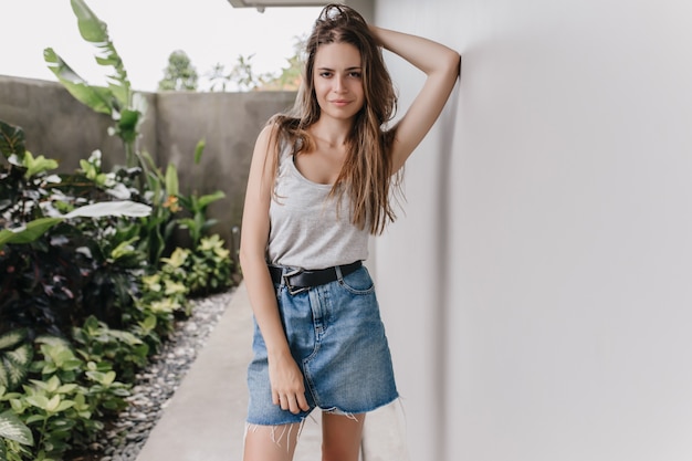 Modelo femenino bien proporcionado en traje de verano de pie en pose de confianza junto a la pared blanca. Foto de chica morena seria lleva falda de mezclilla mirando