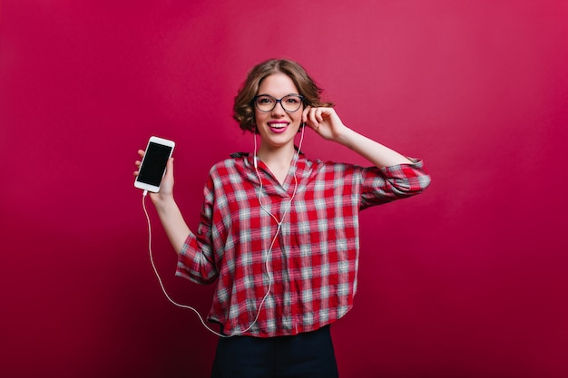 Modelo femenino blanco entusiasta con peinado corto de moda posando con teléfono Chica elegante en camisa a cuadros clarete con smartphone.