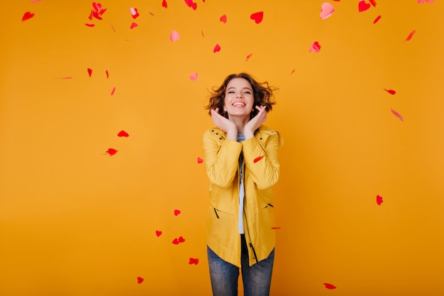 Modelo femenino blanco entusiasta en ropa casual de moda posando con sonrisa feliz. Muchacha maravillosa en la chaqueta amarilla que se coloca bajo corazones caídos.