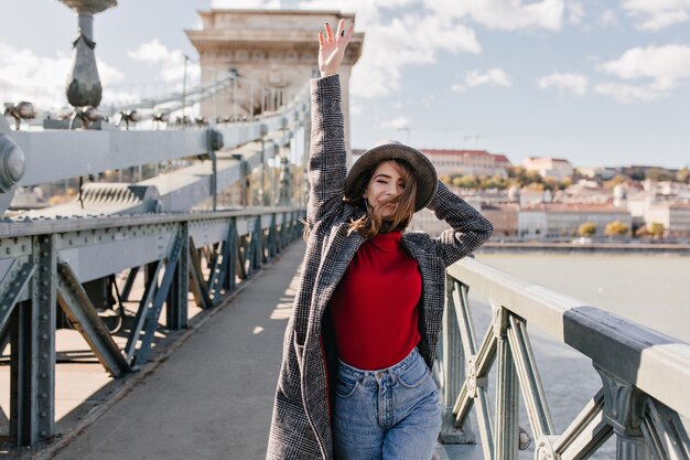 Modelo femenino blanco extático en abrigo de tweed gracioso bailando en el puente sobre el río