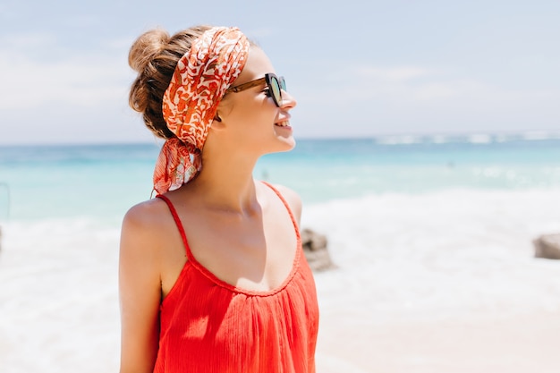Modelo femenino blanco feliz con la presentación de la cinta roja. Tiro al aire libre de elegante chica con estilo en gafas de sol sonriendo durante el paseo por la costa del océano.