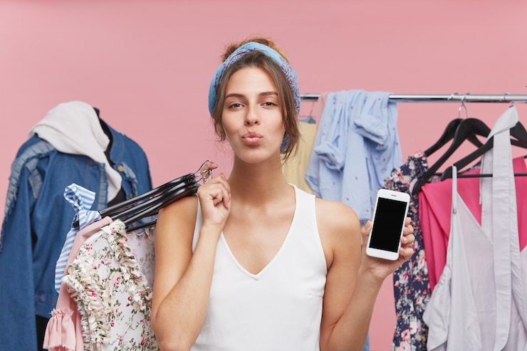 Foto gratuito modelo femenino complacido presionando sus labios, de pie contra manequin y estante con ropa, sosteniendo hagers con ropa y teléfono celular con pantalla en blanco, de buen humor después de una compra exitosa