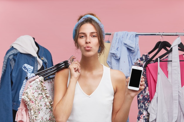 Modelo femenino complacido presionando sus labios, de pie contra manequin y estante con ropa, sosteniendo hagers con ropa y teléfono celular con pantalla en blanco, de buen humor después de una compra exitosa