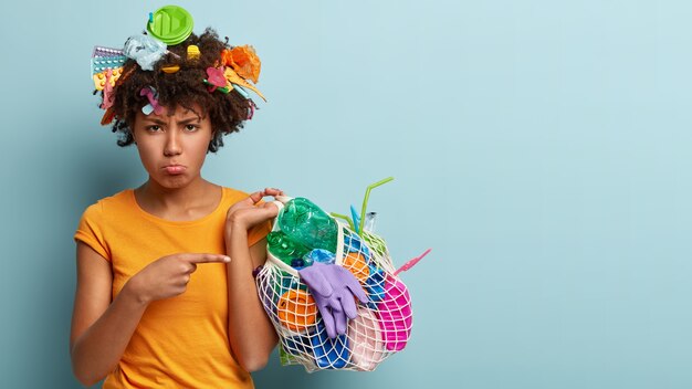 Modelo femenino descontento con piel negra, recoge basura, señala desperdicios de plástico con disgusto, hace trabajo voluntario, protege el medio ambiente, se para sobre una pared azul con espacio libre para su texto