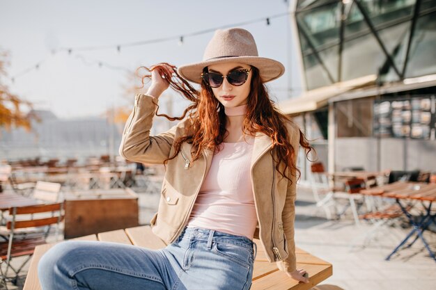 Modelo femenino despreocupado en gafas oscuras posando con sombrero en street cafe