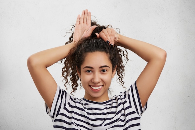 Foto gratuita modelo femenino divertido tiene el pelo nítido, tonto solo, finge ser pequeño conejo o gatito, hace orejas con las manos.