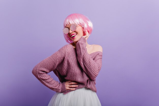 Modelo femenino entusiasta en suéter y falda bailando y sonriendo. Mujer joven inspirada con el pelo rosado aislado en la pared púrpura.