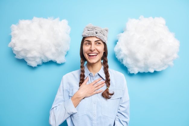 El modelo femenino europeo positivo mantiene la mano en el pecho sonríe felizmente se siente muy feliz mira arriba lleva máscara de dormir y poses de camisa en azul