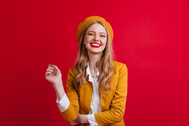 Modelo femenino francés dichoso riendo. Vista frontal de la chica rubia con boina aislada en la pared roja.