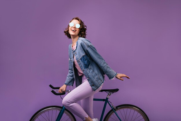 Modelo femenino glamoroso en gafas brillantes posando con bicicleta Chica bastante rizada expresando emociones positivas sobre fondo púrpura