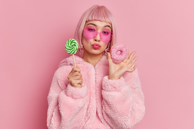 Modelo femenino de glamour de pelo rosa de moda romántica tiene gusto por lo dulce sostiene piruleta y donut glaseado vestido con abrigo de piel cálido gafas de sol en forma de corazón de moda