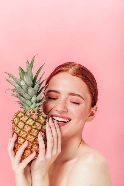 Modelo femenino inspirado con piña. Disparo de estudio de risa chica jengibre con frutas posando sobre fondo rosa.
