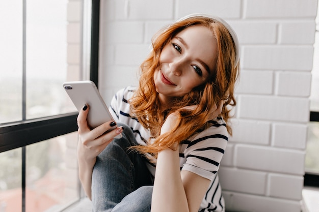 Modelo femenino lindo en auriculares mirando con sonrisa. Atractiva mujer de jengibre con teléfono en mano sentado en casa.