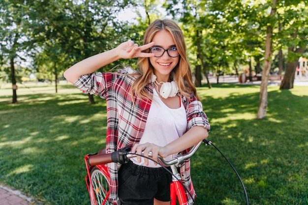 Modelo femenino lindo en vasos posando con el signo de la paz en la naturaleza. Adorable chica rubia montando en bicicleta en el parque.