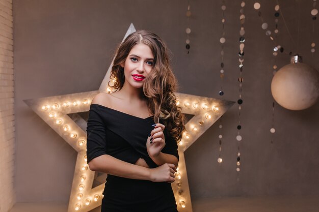 Modelo femenino con maquillaje brillante en vestido negro sonriendo suavemente en la pared gris con estrella ardiente