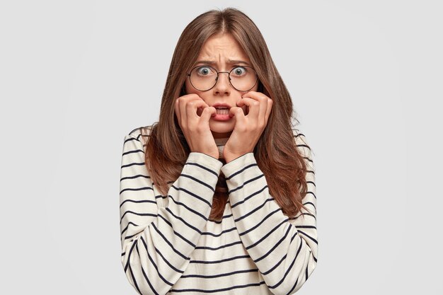 El modelo femenino neurótico asustado mira con ansiedad, siente miedo de algo, vestido con un suéter casual, aislado sobre una pared blanca. Estupendo estudiante teme pasar el examen. Concepto de reacción