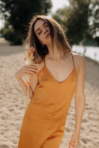 Modelo femenino con peinado oscuro vestido con vestido naranja está posando en la cámara con los ojos cerrados al aire libre en el fondo de la playa de arena