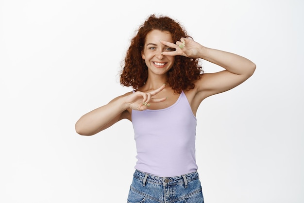 Foto gratuita modelo femenino pelirrojo positivo con cabello rizado que muestra gestos de paz cerca de la cara y sonriendo feliz de pie sobre fondo blanco