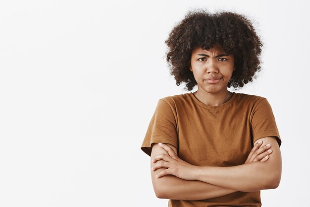 Modelo femenino de piel oscura insatisfecho, intenso y ofendido, disgustado con peinado afro, tomados de la mano cruzados sobre el pecho, frunciendo el ceño y frunciendo los labios, ofendido o decepcionado por la pared gris