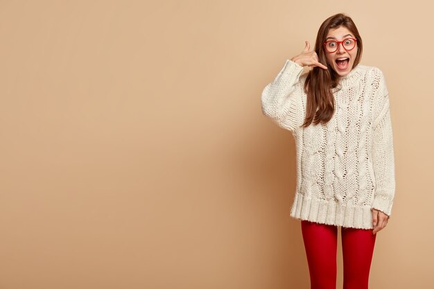 Modelo femenino positivo emocional mantiene los dedos en el gesto de llamarme, usa gafas transparentes, suéter blanco