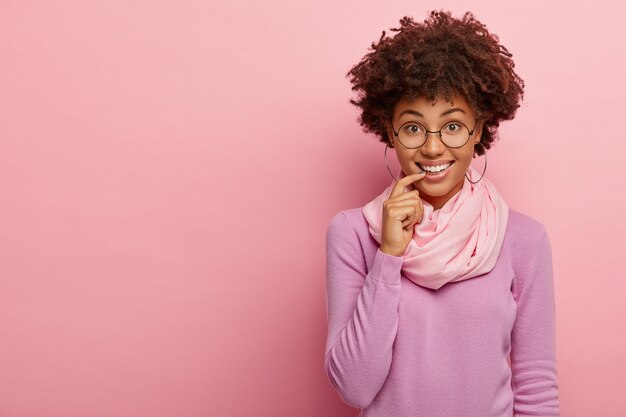 Modelo femenino positivo de piel oscura con una sonrisa encantadora, mantiene el dedo índice en los labios, mira a través de los anteojos, usa modelos de saltadores casuales en interiores, tiene expresión facial relajada