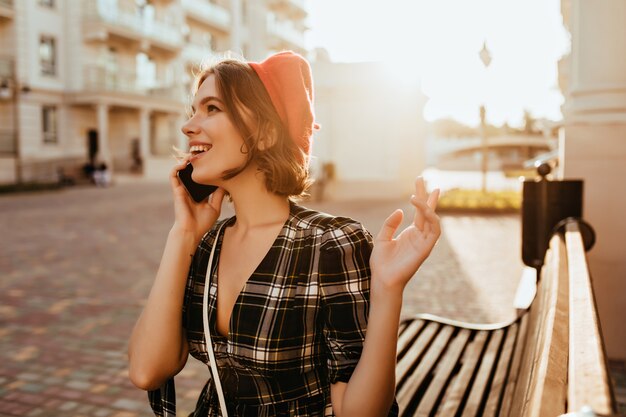 Modelo femenino positivo en ropa elegante disfrutando del día de otoño. Foto exterior de hermosa mujer rizada en boina roja hablando por teléfono.