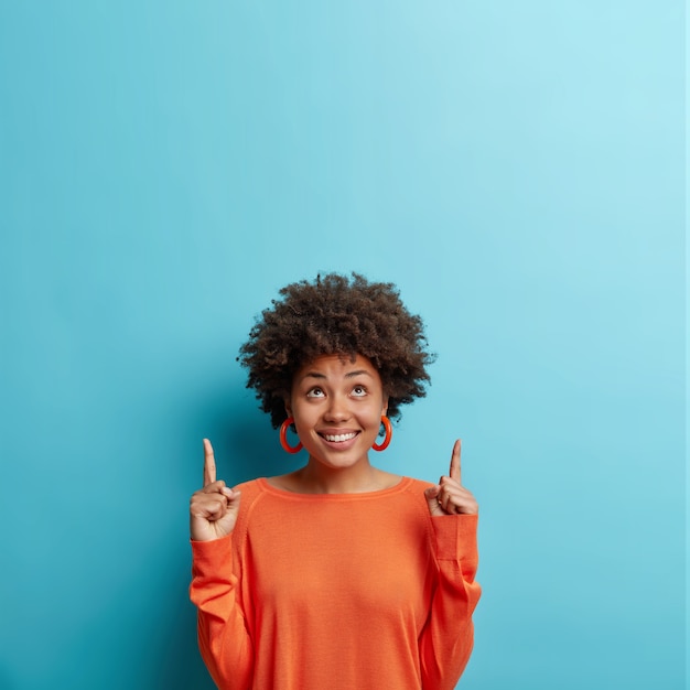 Foto gratuita el modelo femenino positivo viste un puente naranja casual con los dedos índice arriba muestra algo deseable hacia arriba