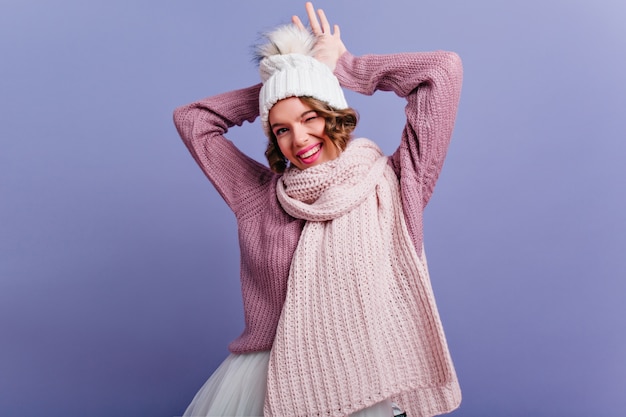 Modelo femenino satisfecho con expresión de cara feliz posando en ropa de invierno y sonriendo. Mujer de pelo corto con bufanda que expresa emociones positivas en la pared púrpura.