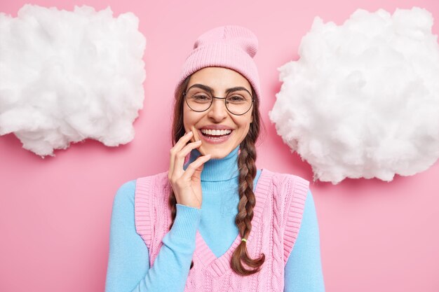 modelo femenino sonríe ampliamente expresa emociones felices tiene dientes blancos viste ropa casual gafas redondas transparentes aisladas en rosa