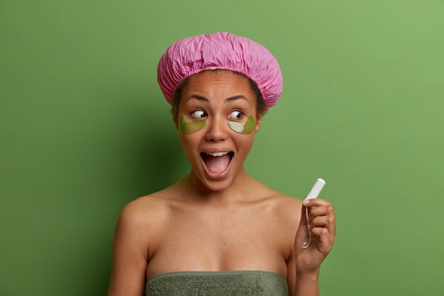 Modelo femenino sonriente saludable envuelto en una toalla de baño, usa parches de belleza verde debajo de los ojos