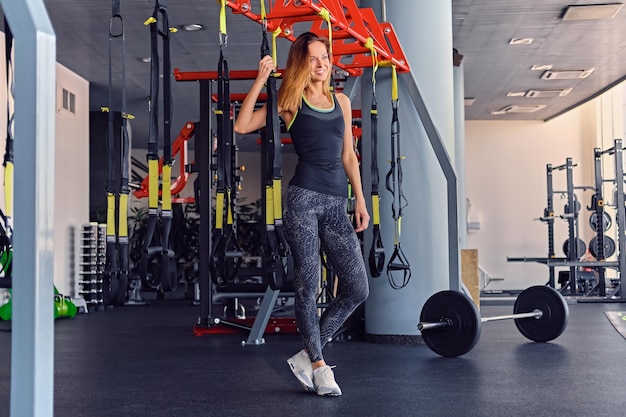 Modelo de fitness femenino delgado atlético en ropa deportiva colorida posando cerca de las tiras de suspensión trx se encuentra en un club de gimnasia.