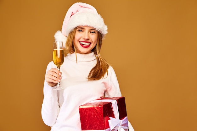 Modelo con gorro de Papá Noel y caja de regalo grande bebiendo champán en la pared de oro