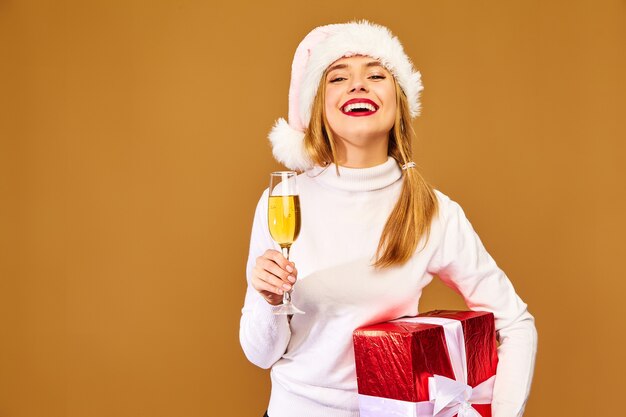 Modelo con gorro de Papá Noel y caja de regalo grande bebiendo champán en la pared de oro