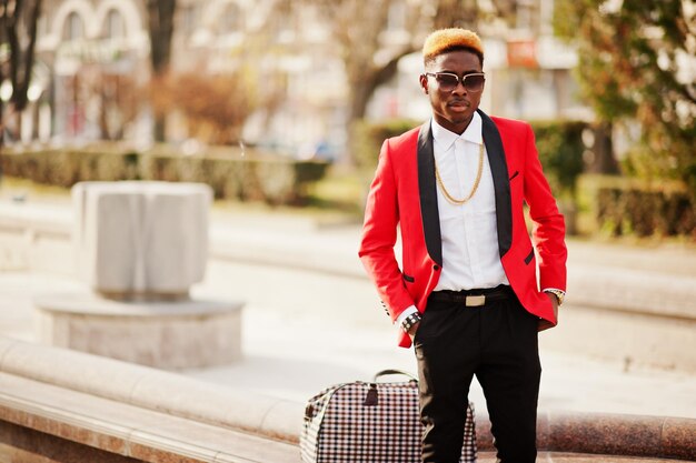 Modelo de hombre afroamericano de moda en traje rojo con cabello destacado y bolso posado en la calle